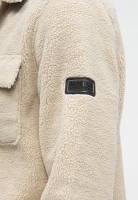 Μπεζ φλις μπουφάν με υφή, μία τσέπη στο στήθος και μαύρο λαστιχένιο λογότυπο στο αριστερό μανίκι.