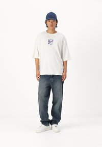 Hvid bomulds t-shirt med en firkantet grafik på brystet, parret med løse blå denim jeans og hvide sneakers. En marineblå kasket fuldender looket.