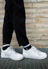 White leather sneakers with perforated toe, round laces, and a raised sole. Worn with black pants and striped socks, standing on a grid surface.