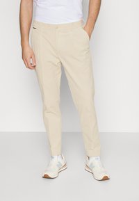 Pantaloni beige con vestibilità su misura, dotati di chiusura a bottone, tasche laterali e una texture liscia. Abbinati a sneakers bianche e blu.