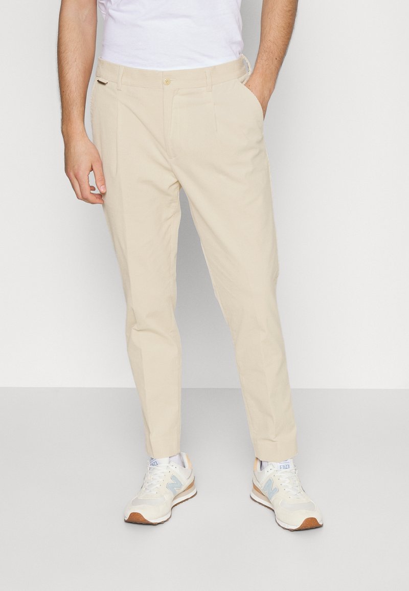 Pantaloni beige con vestibilità su misura, dotati di chiusura a bottone, tasche laterali e una texture liscia. Abbinati a sneakers bianche e blu.