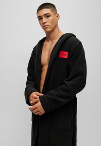 Black hooded bathrobe in towel material, featuring a front pocket and a red branded patch labeled "HUGO." Open front design, medium length.