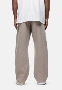 Man in een losse beige broek, witte sneakers en een wit T-shirt, gezien van achteren tegen een effen achtergrond.