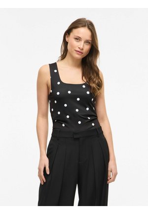 Femme aux longs cheveux bruns portant un haut noir sans manches à pois blancs et un pantalon noir plissé taille haute.