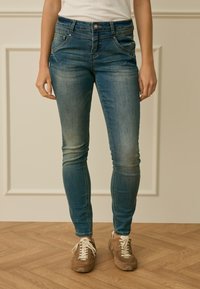 Osoba stojící na dřevěné podlaze před béžovým obložením stěny, oblečená v vybledlých modrých úzkých džínách a hnědých teniskách.