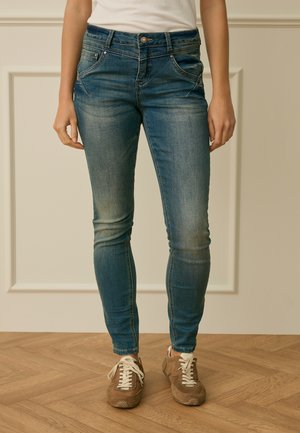 Personne portant un jean skinny bleu délavé et des baskets marron, debout sur un sol en bois devant un panneau mural beige.