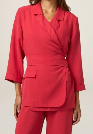 Femme portant un blazer rouge croisé à manches trois-quarts et un pantalon rouge assorti sur un fond uni.