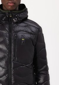 Giacca a vento nera imbottita con chiusura zip frontale, colletto alto e taschino sul petto. Presenta una texture lucida e trapuntata con un'etichetta del logo.