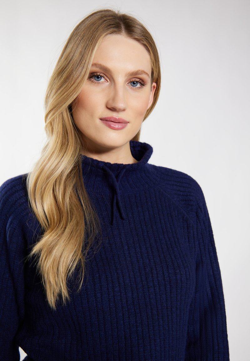 Donna bionda con lunghi capelli che indossa un maglione a coste blu navy con colletto alto, che guarda leggermente oltre la macchina fotografica su uno sfondo semplice.