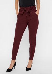 Pantalon taille haute bordeaux avec détail noué, doté de poches latérales et d'une coupe fuselée, associé à des chaussures à talons noires.