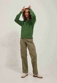 Pulover verde tricotat cu un design cu nervuri, asortat cu pantaloni cargo de culoare măslinie deschisă, care au buzunare laterale. Adidași albi completează ținuta.