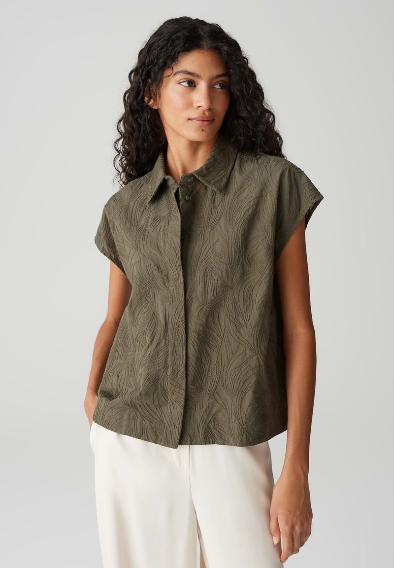 Olijfgroene shirt met korte mouwen, voorzien van een getextureerd bloemenpatroon, met kraag en knoopsluiting aan de voorkant, gecombineerd met lichtgekleurde broeken.