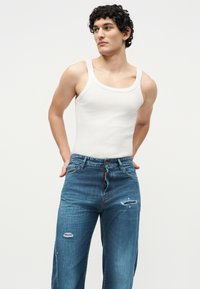 Joven con pelo corto y rizado, vestido con una camiseta blanca sin mangas y vaqueros azules desgastados, de pie con las manos detrás de la espalda.