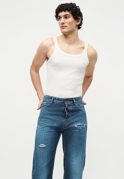 Νέος άνδρας με κοντά σγουρά μαλλιά, που φοράει λευκό φανελάκι και μπλε τζιν με φθορές, στέκεται με τα χέρια πίσω από την πλάτη.