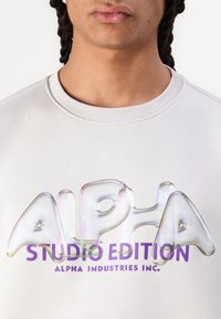 Sweatshirt gris clair avec un grand graphique "ALPHA" brillant et multicolore avec un effet 3D et le texte "STUDIO EDITION" en violet en dessous.