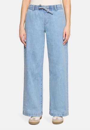 Jasnoniebieskie szerokie dżinsy o luźnym kroju z elastyczną talią i sznurkiem, noszone z białymi trampkami.