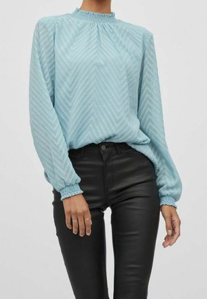 Lichtblauwe blouse met een geplooide hals en lange mouwen, voorzien van een chevronpatroon en afgewerkte manchetten, gecombineerd met zwarte, aansluitende jeans.