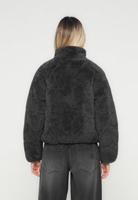 Kvinde med skulderlangt ombre hår iført en mørkegrå tekstureret fleecejakke og sorte jeans, vist fra ryggen.