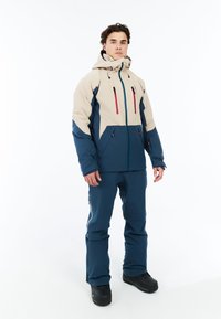 Heren tweekleurige jas in beige en donkerblauw met ritszakken en verstelbare capuchon, gecombineerd met bijpassende blauwe broek en zwarte laarzen.