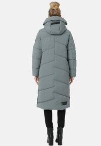 Abrigo acolchado largo gris con capucha, que presenta patrones de acolchado diagonales y un parche con el logo. Combinado con pantalones negros y botas hasta la rodilla.