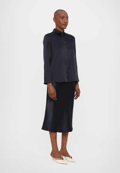 Navy button-up shirt with a collar, paired with a smooth, knee-length navy skirt. The model wears cream slip-on heels. Smooth texture throughout.