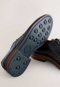 Black leather dress shoe with decorative broguing, featuring a navy rubber sole with circular traction points and a wood-stacked heel.