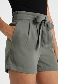 Zelené šortky s vysokým staženým pasem a odnímatelným pásem s mašlí. Mají boční kapsy a hladkou texturu látky.