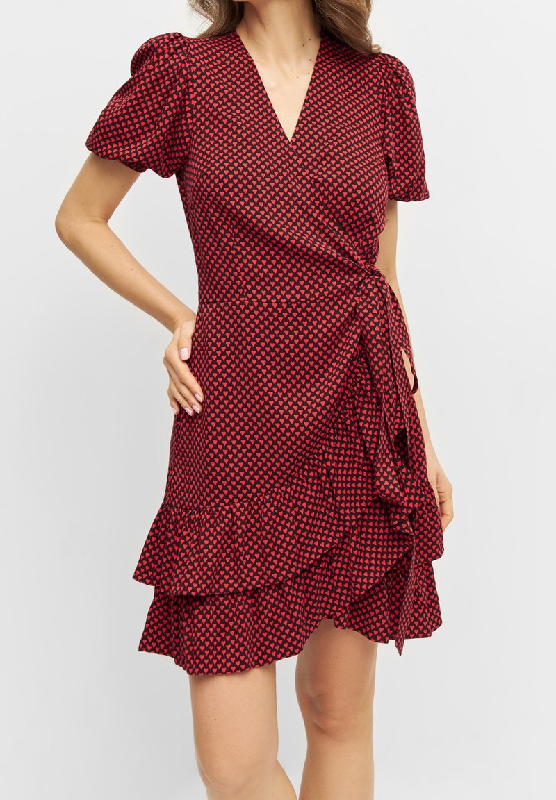 Robe rouge à manches courtes bouffantes, motif de cœurs dispersés, décolleté en V, ourlet volanté et détail de taille à nouer. Texture de tissu lisse.