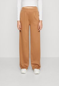 Pantalons larges beiges avec des rayures blanches verticales, dotés d'une taille élastique haute. Portés avec un haut ajusté à manches longues blanc et des baskets blanches.