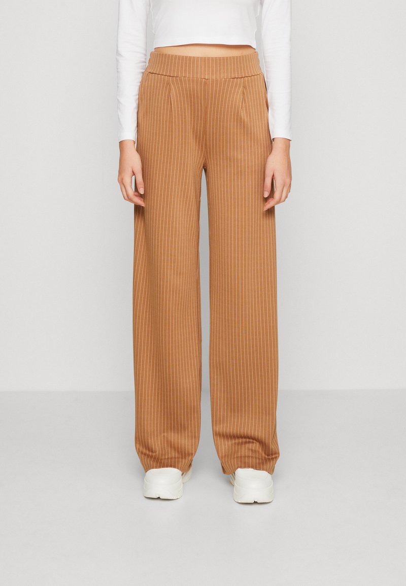 Pantalons larges beiges avec des rayures blanches verticales, dotés d'une taille élastique haute. Portés avec un haut ajusté à manches longues blanc et des baskets blanches.