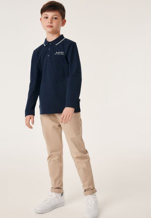 Marineblå langærmet polo med hvid krave og logo, beige tætsiddende bukser og hvide sneakers, stående på en neutral baggrund.
