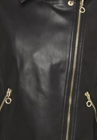 Veste noire en similicuir avec trois fermetures éclair dorées, une fermeture éclair verticale au centre et deux fermetures éclair diagonales sur les poches latérales munies de tirettes circulaires.