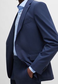 Chaqueta de traje azul marino con acabado texturizado, un bolsillo en el pecho y corte entallado. Incluye una camisa de botones azul claro debajo.