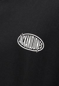 Λευκό κείμενο "JACK AND JONES" με αστέρια μέσα σε οβάλ στο στήθος από μαύρο ύφασμα, πιθανώς μπλουζάκι ή φούτερ.