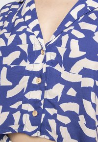 Blusa di cotone con sfondo blu e motivi astratti bianchi, colletto a punte e bottoni in legno sul davanti.