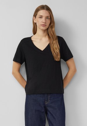 Frau mit langen hellbraunen Haaren steht mit den Händen hinter dem Rücken, trägt ein schwarzes T-Shirt mit V-Ausschnitt und kurzen Ärmeln sowie dunkelblaue Jeans vor grauem Hintergrund.