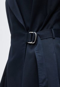 Κοντινό πλάνο σε σκούρο μπλε παλτό τρεντς με υφασμάτινη ζώνη που στερεώνεται με ασημένια αγκράφα διπλού D στο ύψος της μέσης.