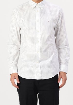 Homme portant une chemise blanche à petits pois noirs et un pantalon noir, debout les mains détendues le long du corps.