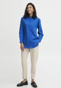 Blue button-up shirt with long sleeves, paired with beige tapered pants and black loafers. Smooth fabric and classic collar design.