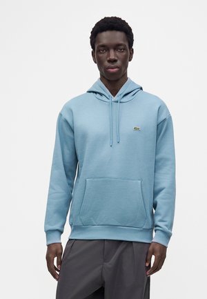 Jeune homme portant un sweat à capuche bleu clair avec poche avant et petit logo vert sur la poitrine gauche sur un fond uni.