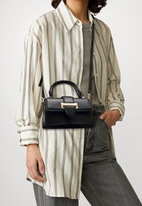 Person som bär en beige och svart randig skjorta, grå jeans, och håller en svart läderhandväska med guldspänne på en axelrem.