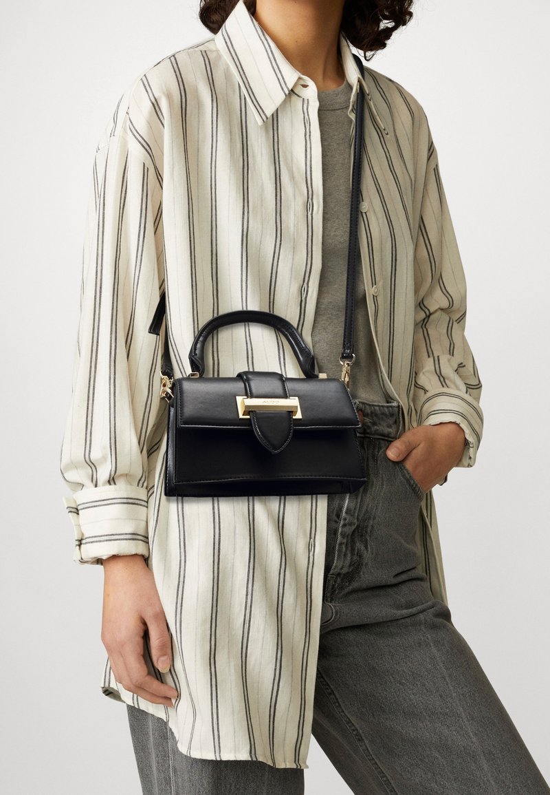 Person som bär en beige och svart randig skjorta, grå jeans, och håller en svart läderhandväska med guldspänne på en axelrem.