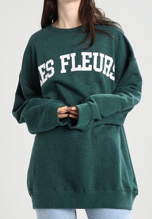 Person wearing oversized dark green sweatshirt with white "LES FLEURS" text, hands clasped at waist, paired with light blue jeans.