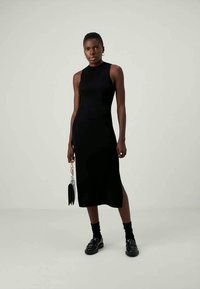 Nainen, joka on pukeutunut mustaan hihattomaan midi-mekkoon, mustiin sukkiin ja mustiin loafer-kenkiin, kantaa pientä mustaa käsilaukkua, jossa on ketjuhihna, seisten yksinkertaista taustaa vasten.