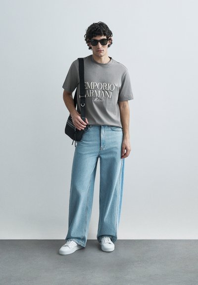 T-shirt cinzenta de algodão com o logótipo "Emporio Armani", jeans de perna larga azul clara, mala preta de tiracolo e sapatilhas brancas. Cabelo encaracolado e óculos de sol.