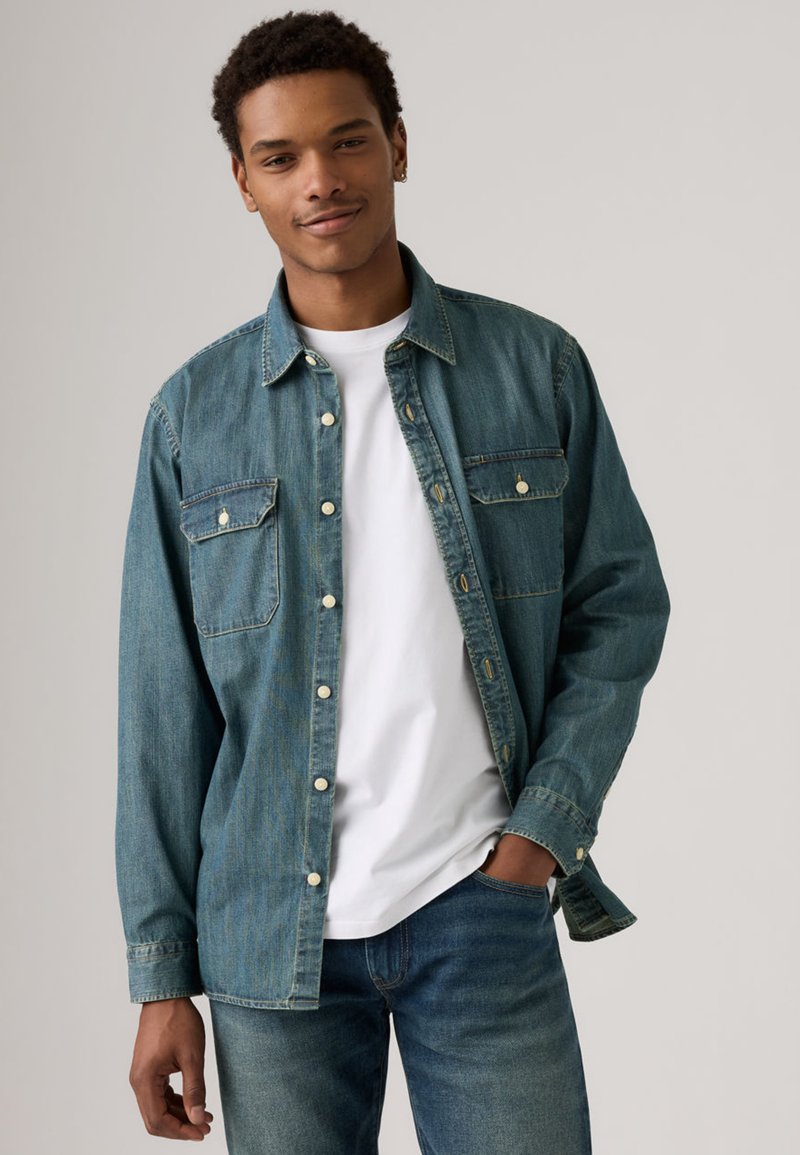 Camicia di denim blu medio, con chiusura frontale a bottoni, due tasche sul petto e colletto classico. Indossata sopra una t-shirt bianca.