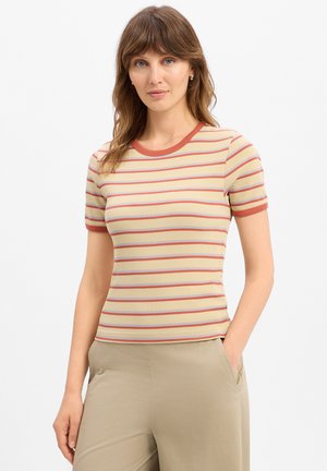 Vrouw met schouderlang bruin haar, die een beige en roestkleurig gestreept T-shirt met korte mouwen en lichtbruine broek draagt, met één hand in de zak.