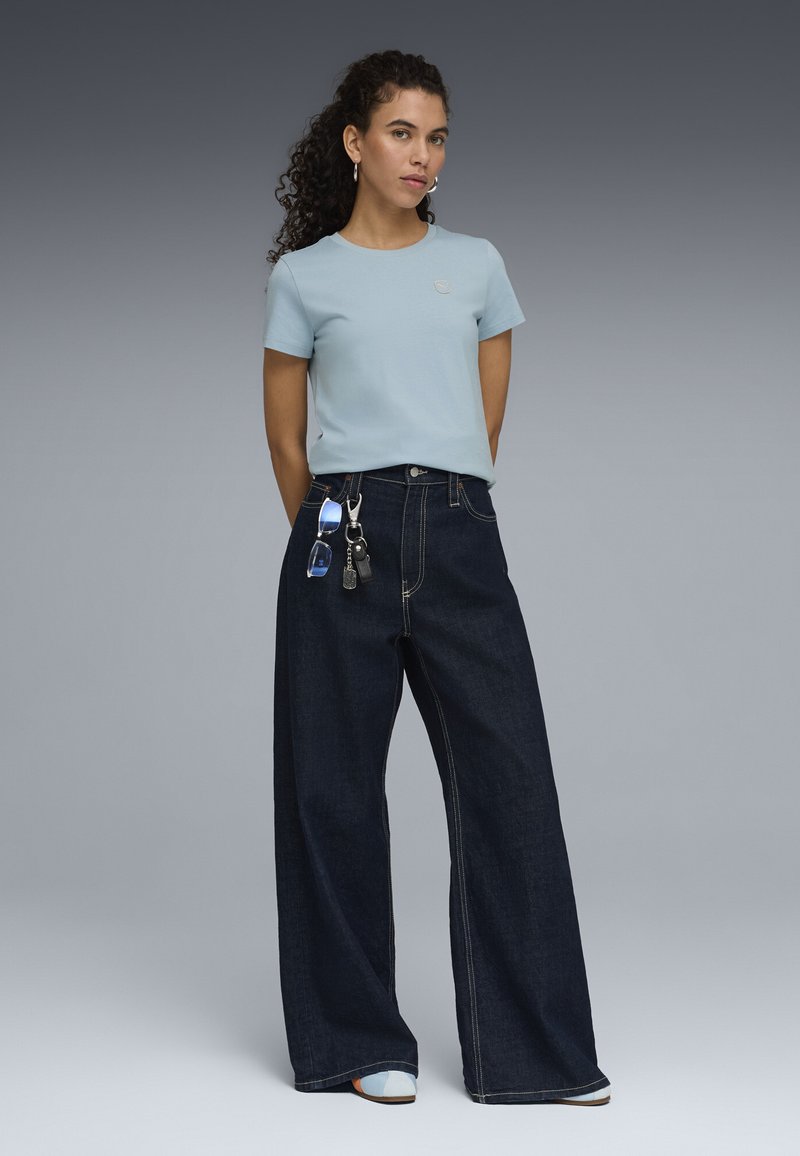 Žena s kučeravými vlasmi, oblečená v svetlomodrom tričku a tmavých džínsach s širokými nohavicami, s okuliarmi na slnko a kľúčmi zavesenými na opaskovom púzdre, stojaca pred sivým pozadím.