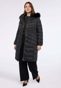 Černá péřová bunda s kapucí s umělým kožešinovým lemováním, prošívaná textura a zlaté zipy. Nositelná přes tmavý top a černé kalhoty, spolu s vysokými podpatky.