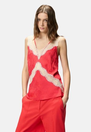 Giovane donna con capelli castani lisci indossa un camisolino rosso in raso con rifiniture in pizzo bianco e pantaloni rossi a gamba larga abbinati, mani nelle tasche.